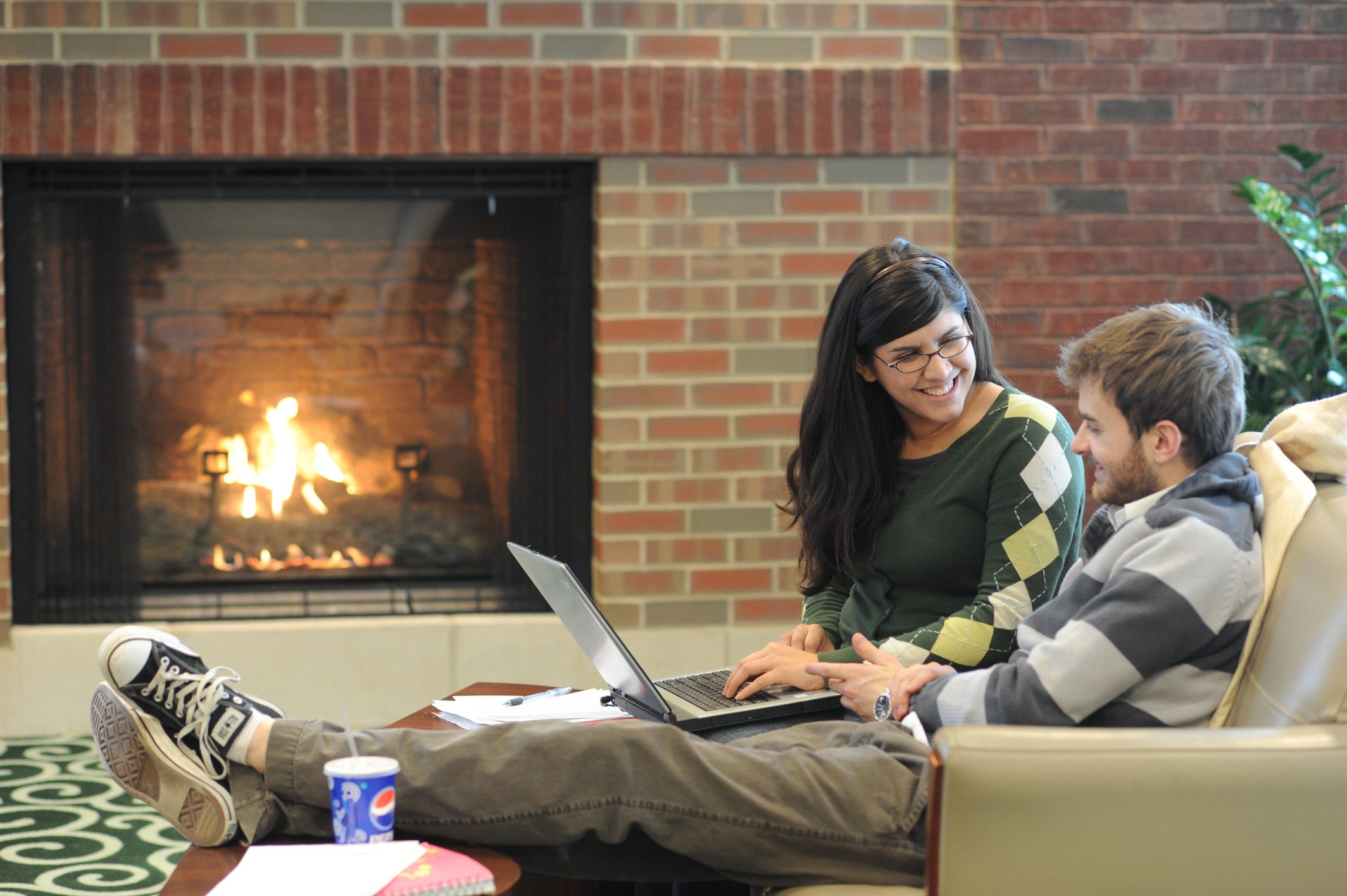 Two people smile at each other as they work on a computer in front of a crackling fireplace