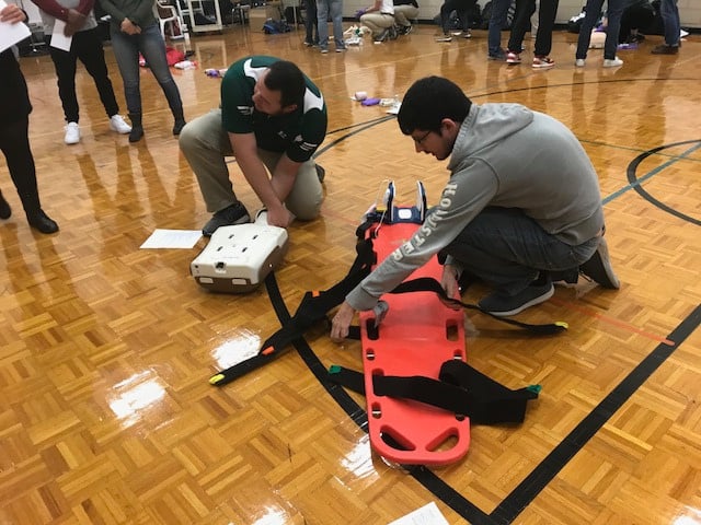 Two participants set up a stretcher and a CPR dummy on a gymnasium floor.