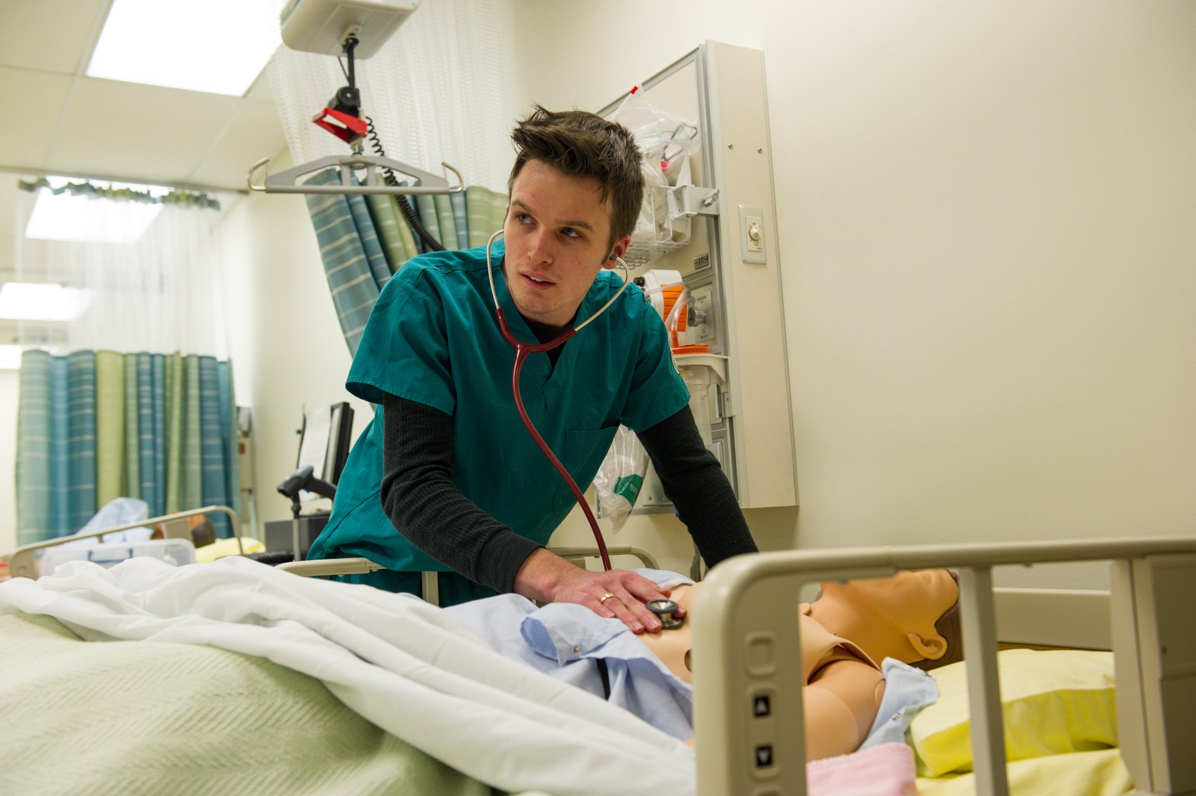 A nursing student in teal scrubs holds a stethoscope to a dummy