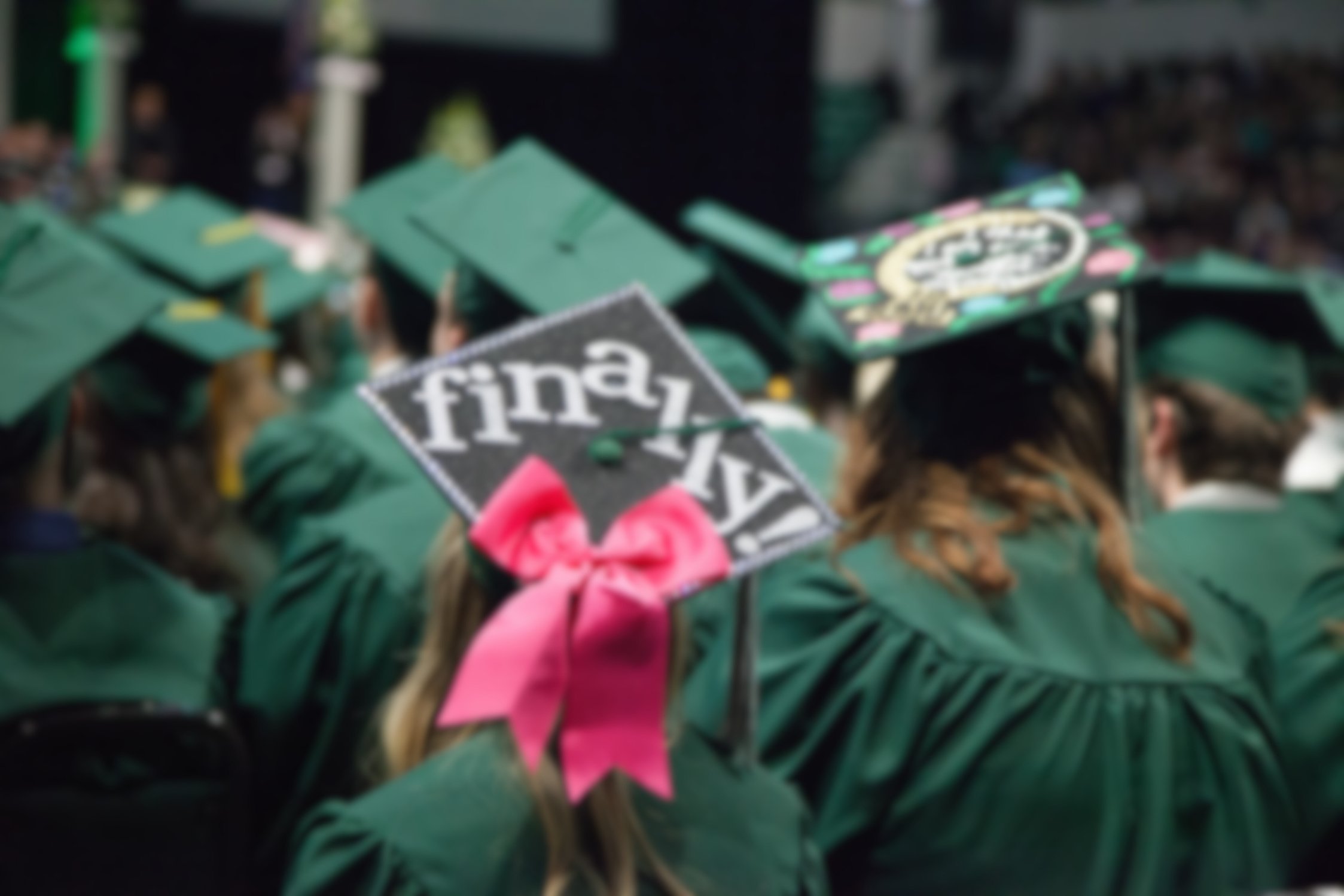 A photo of graduates in green caps and gowns at a graduation ceremony, one of the caps spells "finally!" 