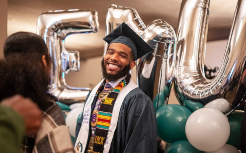 An EMU graduate in a graduation cap and gown smiles at the camera while standing in front of silver balloons that spell "EMU"