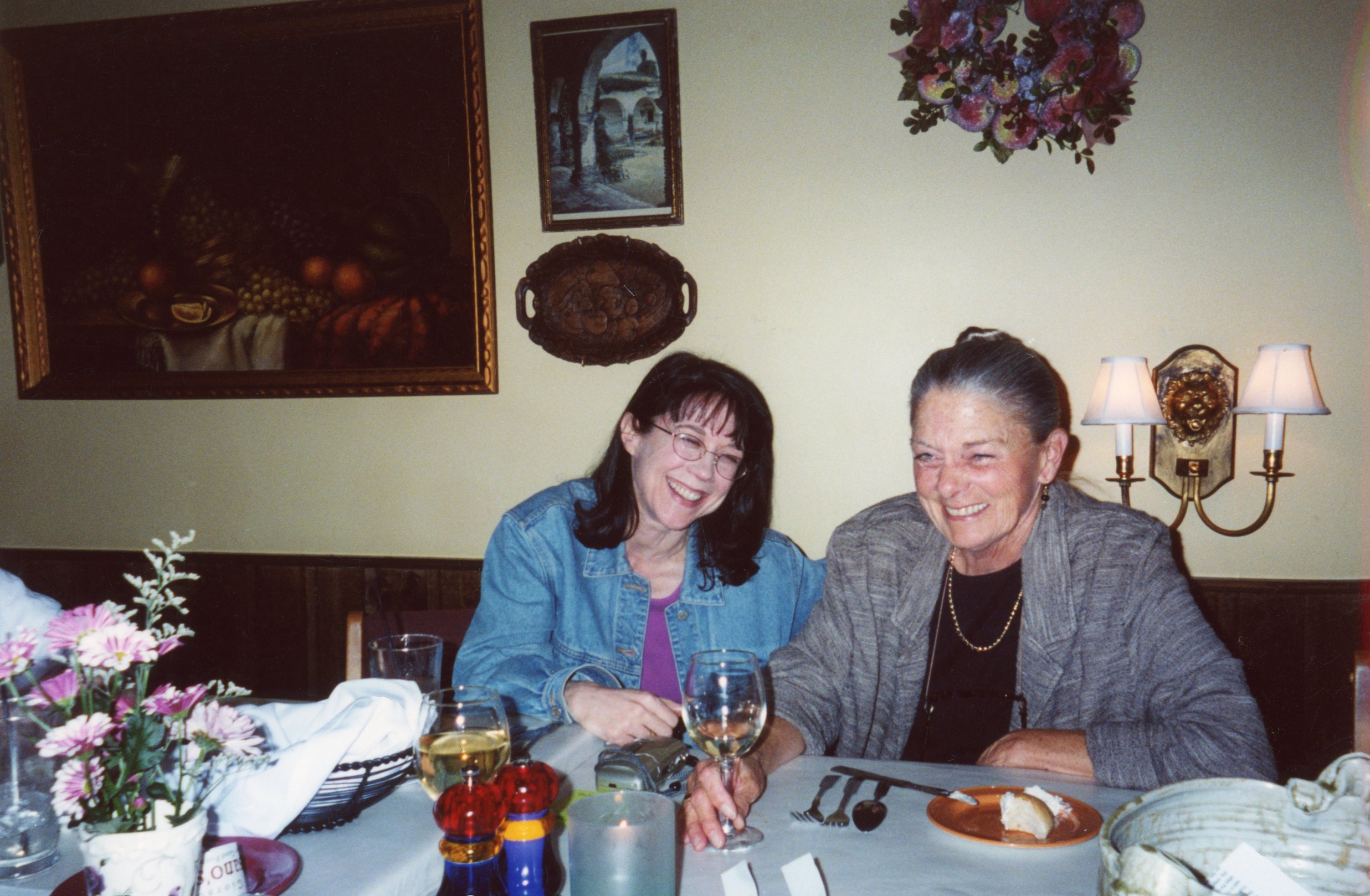 Annette Martin laughs with a friend at a table with wine glasses and a painting on a wall