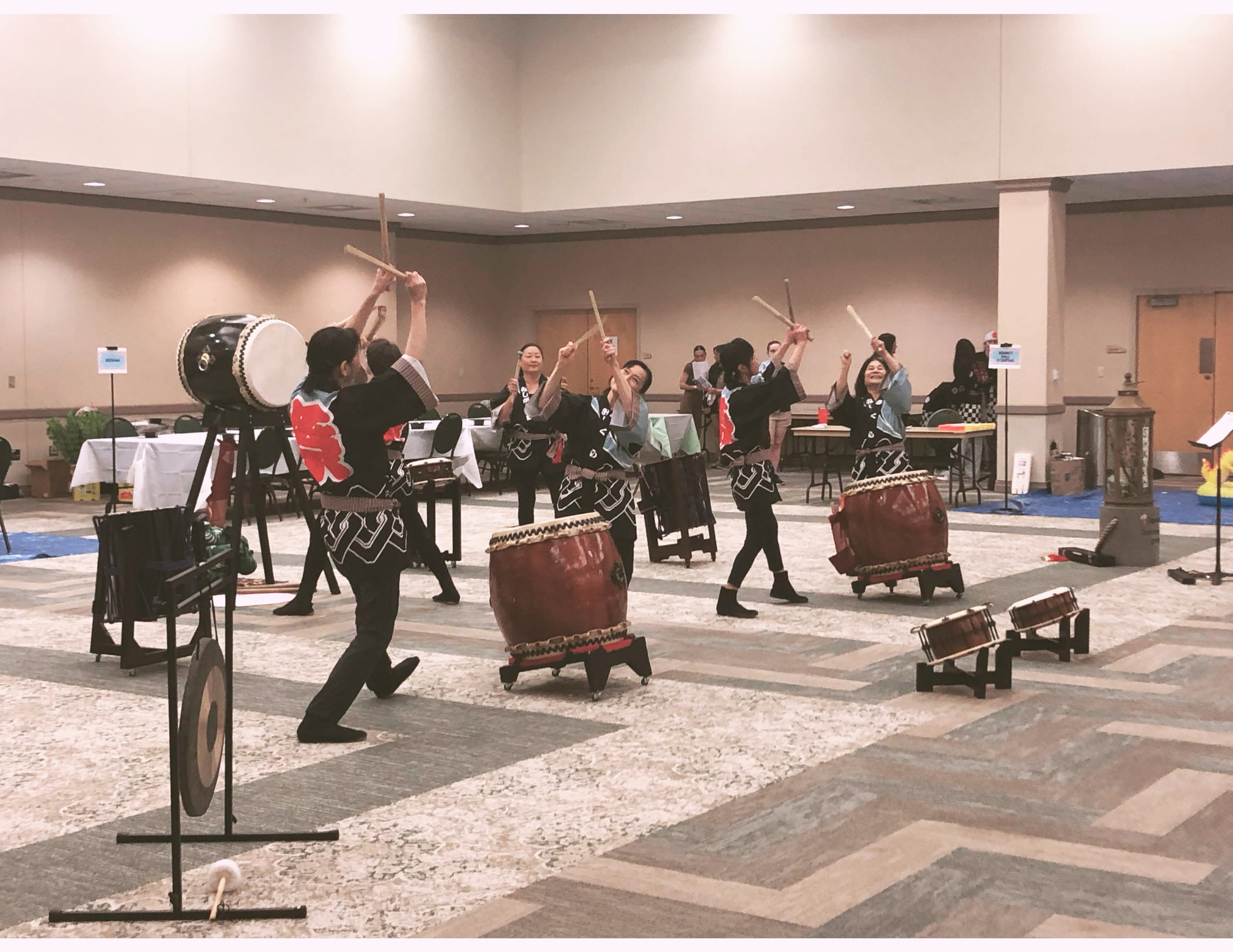 a group of people dressed in black, white, and red traditional Japanese garb playing Japanese drums (taiko) in a campus conference hall