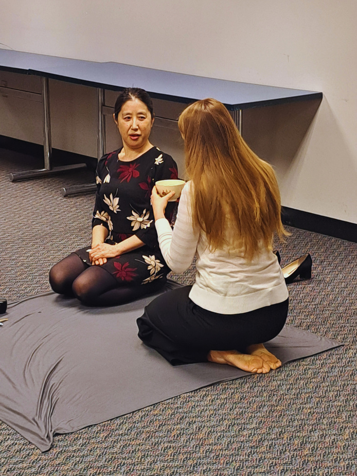 Two people kneel on the floor while one holds a Japanese tea cup. 