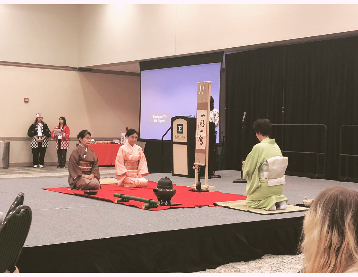 two people in kimono perform a tea ceremony while sitting on a red carpet in front of an audience