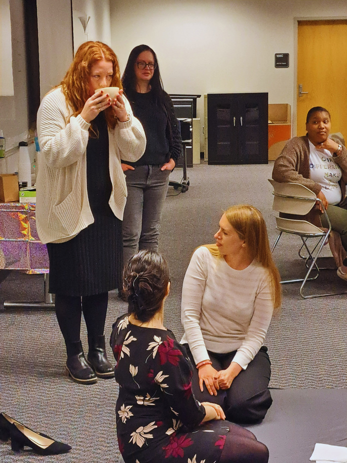 People kneel and stand in a conference room while one person sips tea from a Japanese tea cup