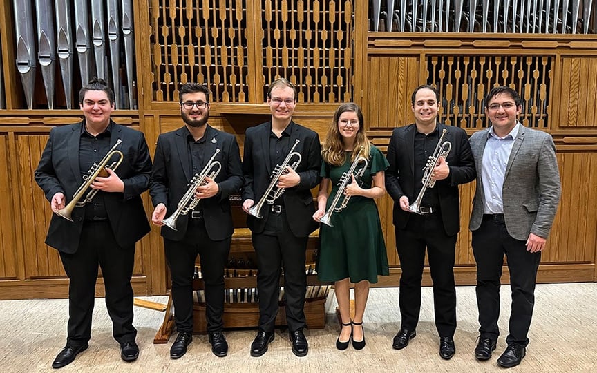 The trumpet quintet, dressed in all black, and their instructor, dressed in a suit, posing for a photo with their trumpets in front of an organ