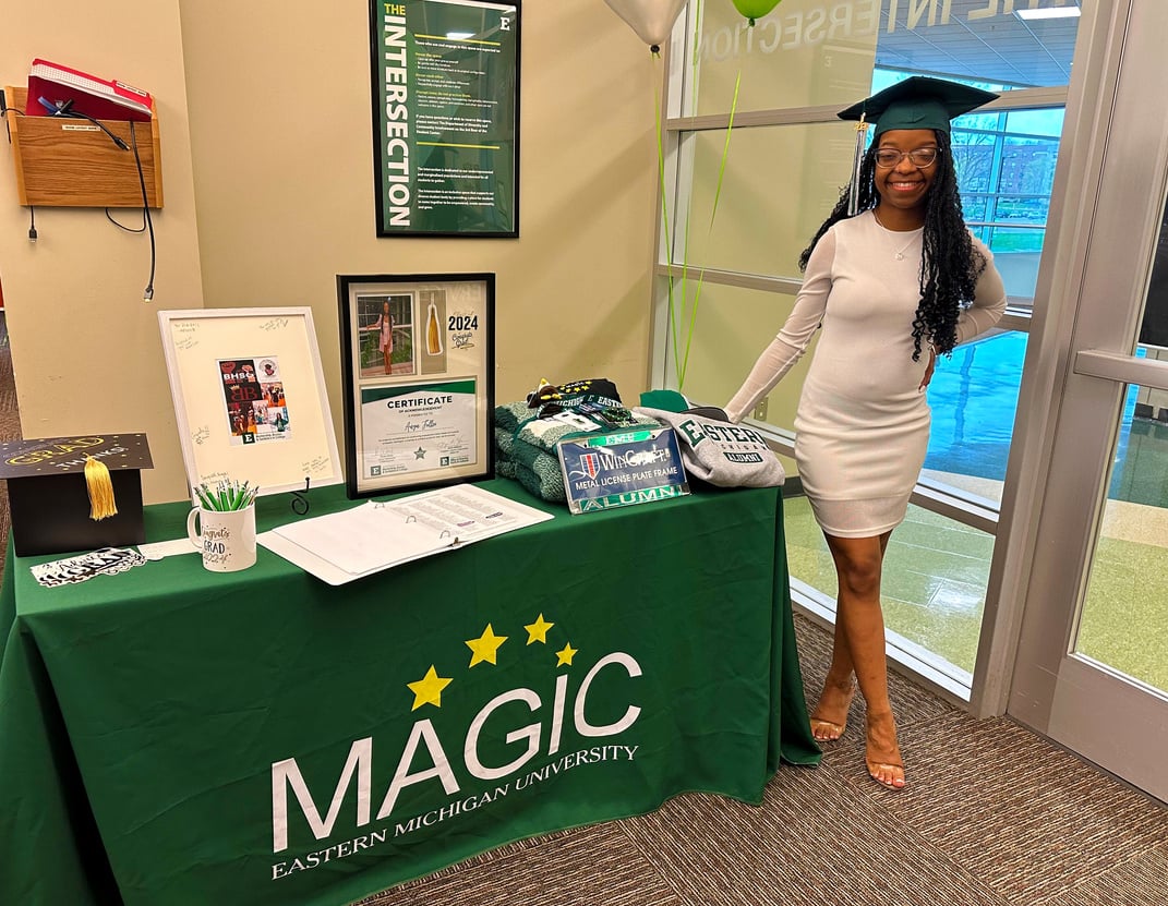 Anya Fuller in a white dress and green graduation cap standing next to a table with a magic sign on it