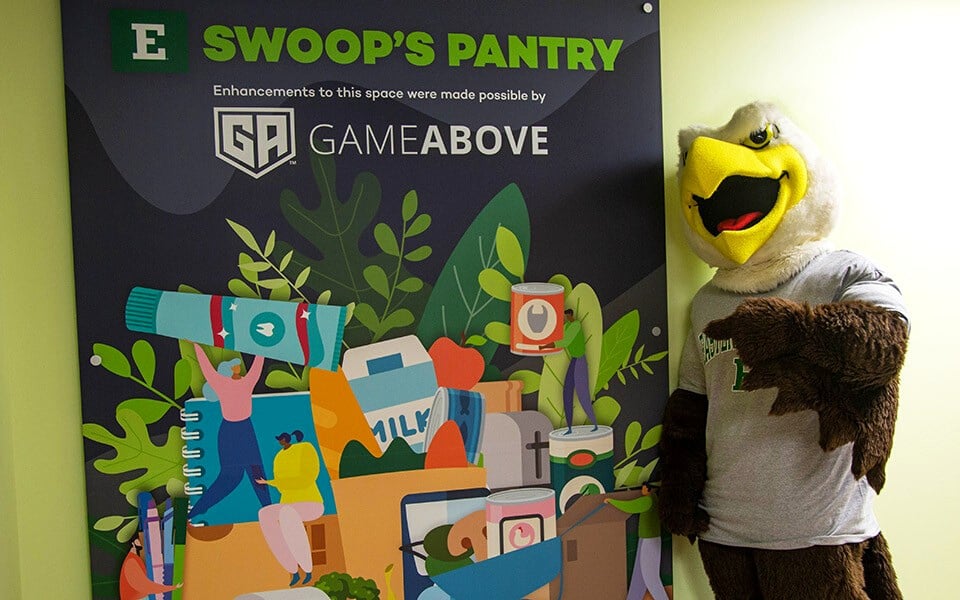 The EMU Eagle mascot stands beside a colorful sign announcing the Swoop's Pantry renovation.