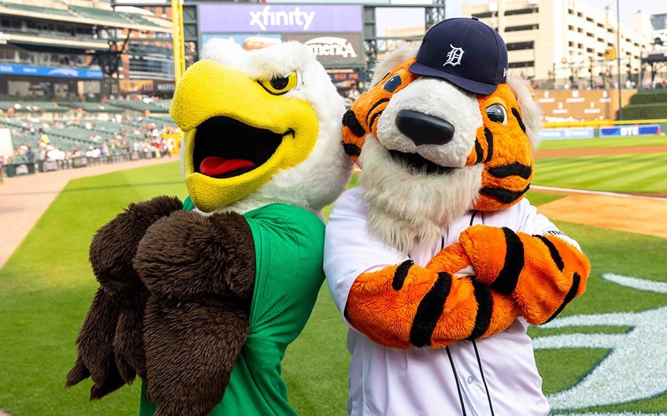 Swoop, the eagle mascot for EMU, and Paws, the Detroit Tigers Baseball mascot, stand back to back in front of Comerica Park baseball stadium