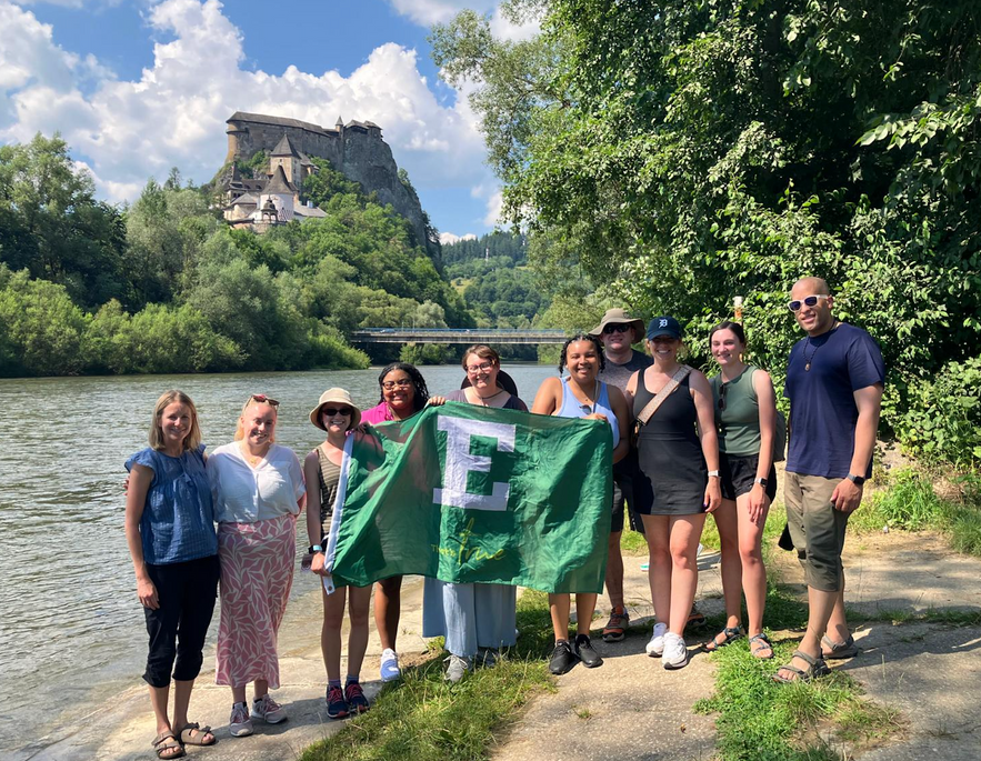 a group of people standing in front of a river with a green EMU flag