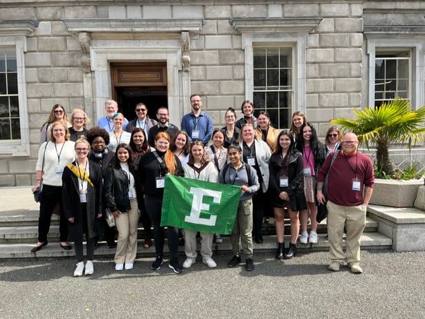 A group photo of students and faculty standing outside of a white brick building with the EMU flag held before them