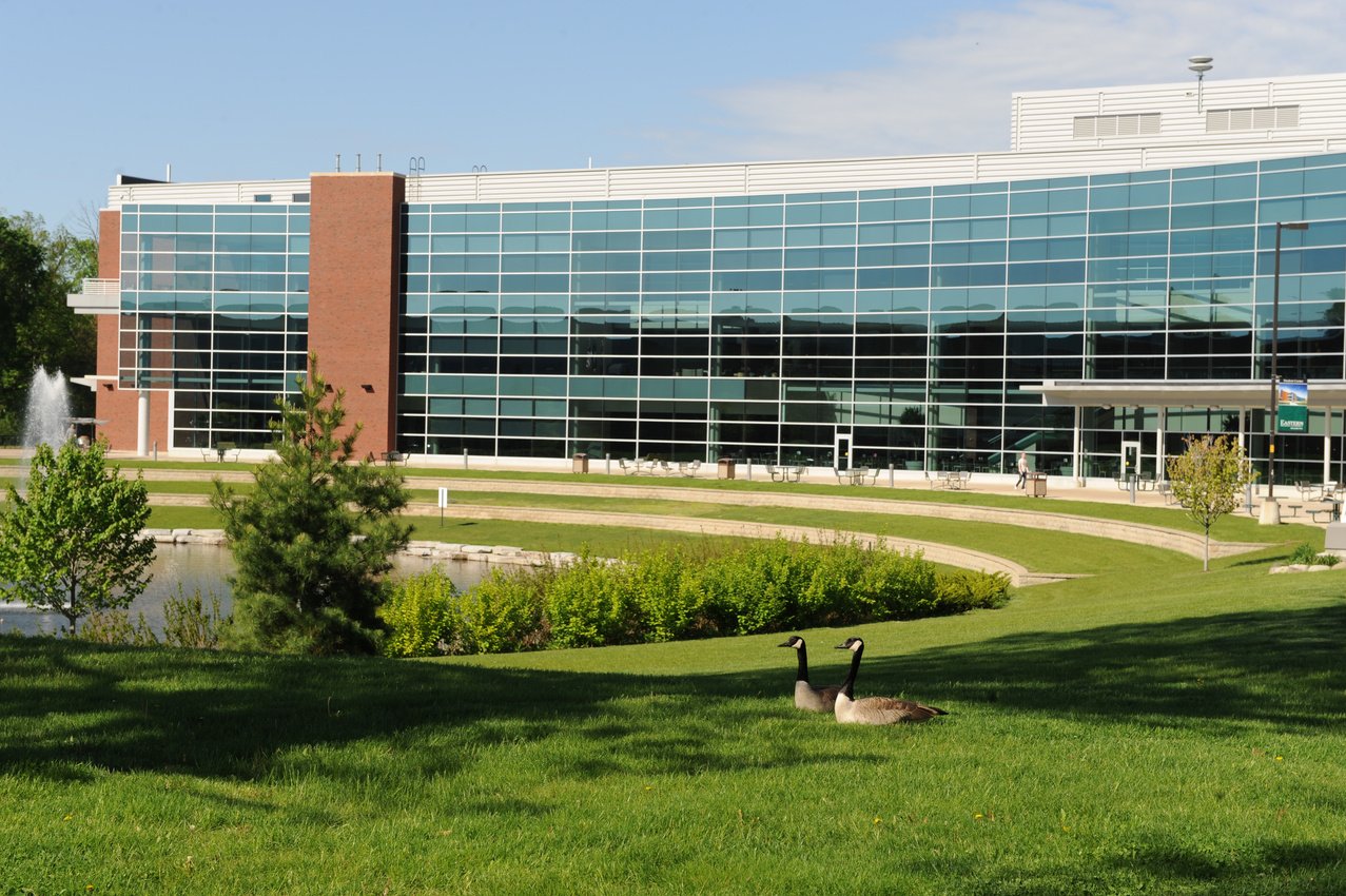 A view of the EMU Student Center from across the nearby pond and green lawn. Two geese sit in the foreground. 