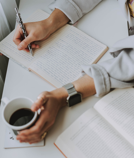 hands writing in an open book and holding a cup of coffee